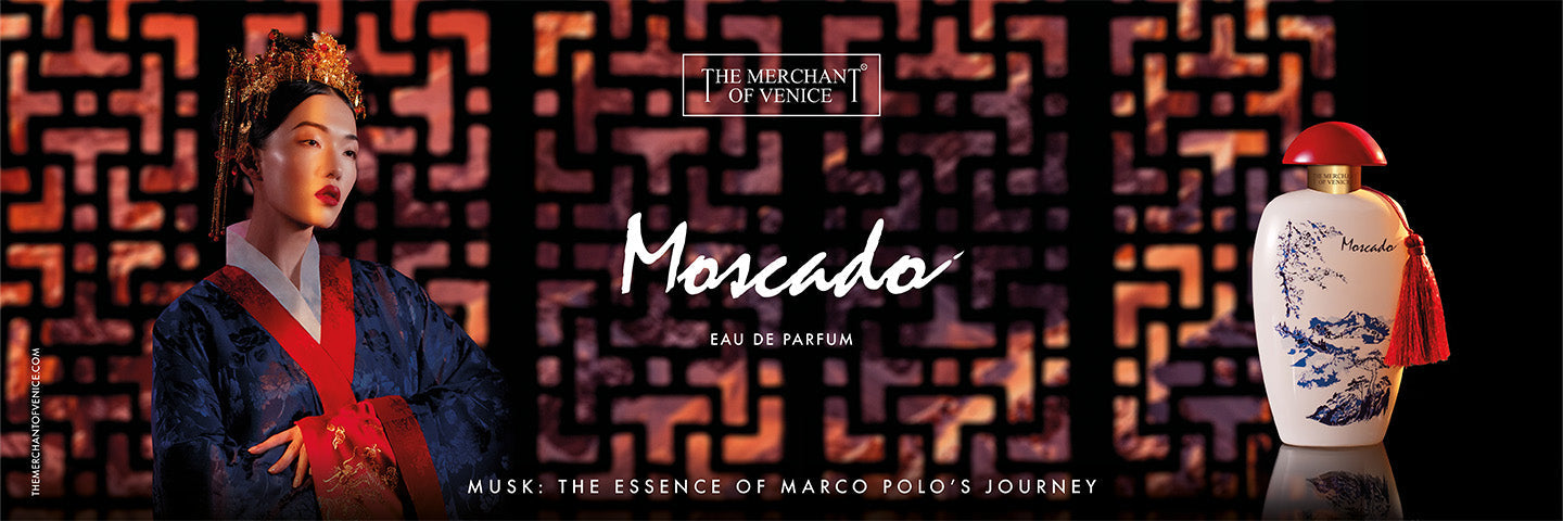 Una donna in abito tradizionale orientale posa accanto al flacone del profumo "Moscado" di The Merchant of Venice. Sullo sfondo, un pannello geometrico nei toni caldi del rosso e nero avvolge la scena in un’atmosfera raffinata ed evocativa.